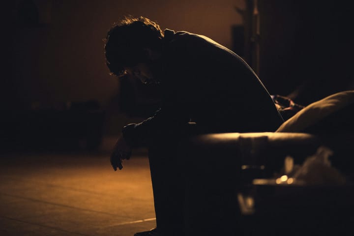 A masc person sits outside stooped over in sadness. They are in full shadow.