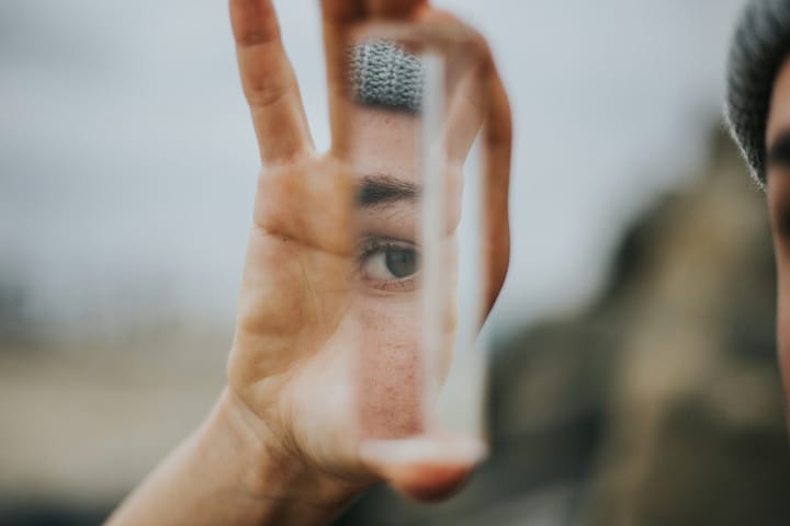 Someone mostly off camera with a beanie holds up a rectangular sliver of mirror. Their eye, part of their brow, and beanie is visible in it
